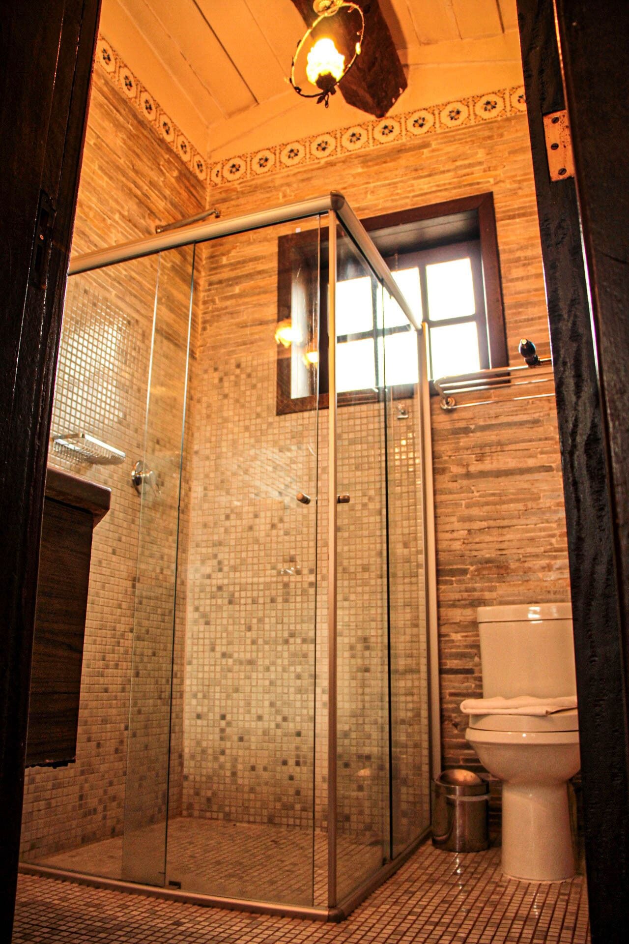Ducha con cerramiento de vidrio, pared de azulejos y ventana sobre el inodoro.