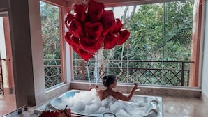 Une femme dans une baignoire remplie de mousse, avec un bouquet de roses rouges et un verre de vin posés sur le bord.
