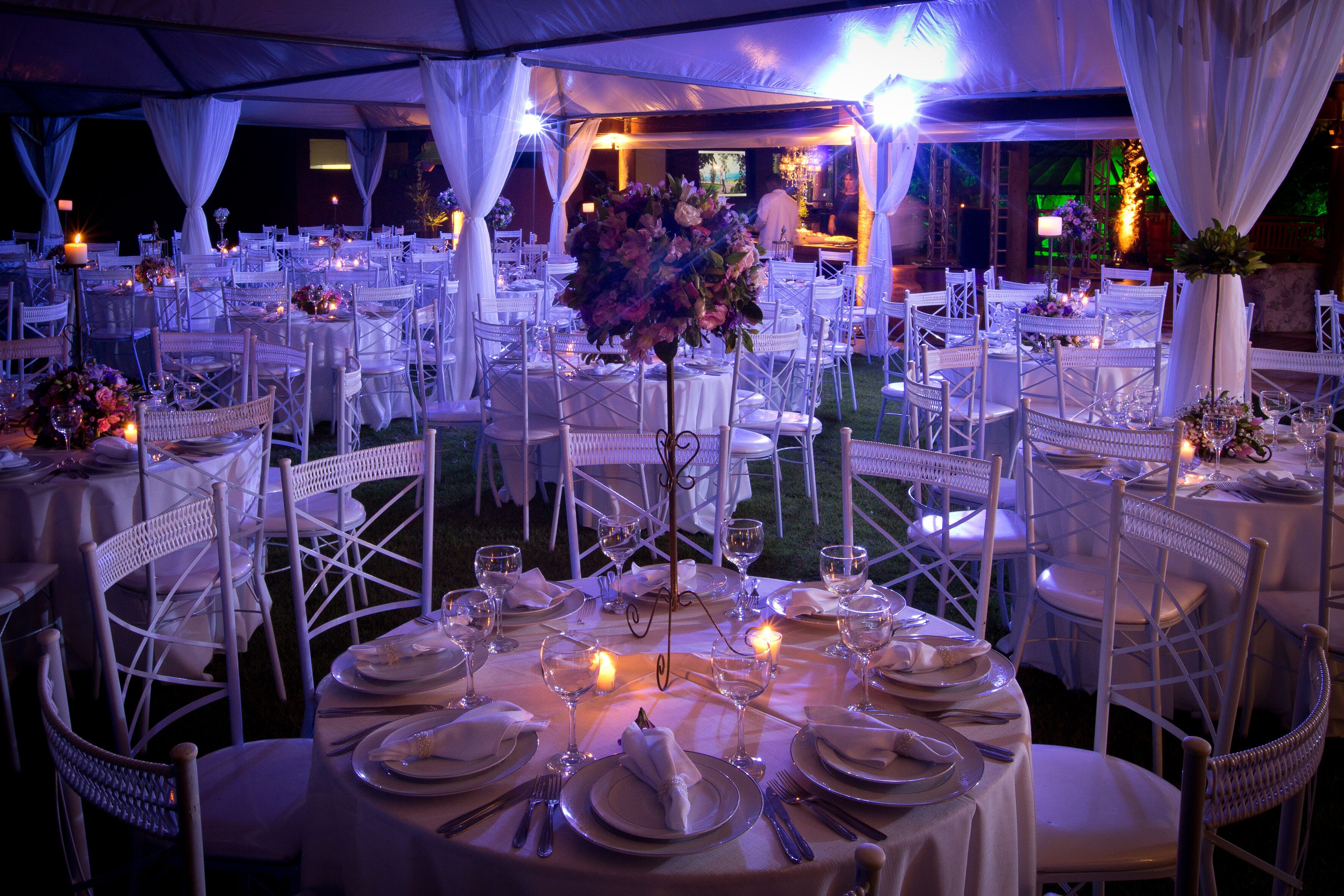 Un área de comedor al aire libre con carpas, mesas redondas, sillas blancas y centros de mesa iluminados con velas.