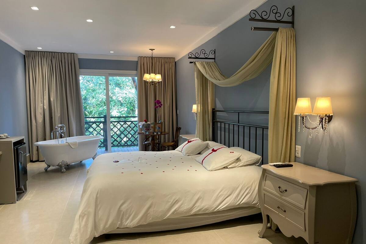 A bedroom with a large bed, a bathtub, a vanity, and a balcony with curtains.