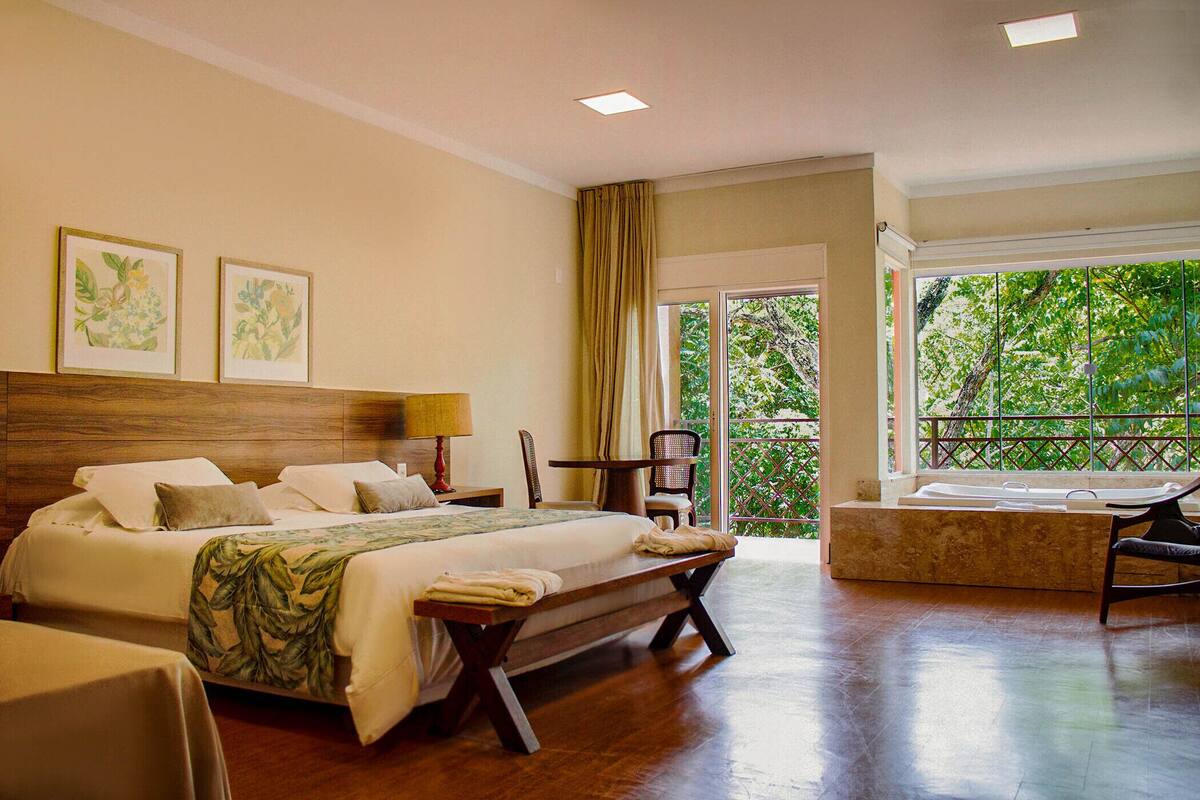 A hotel room with a large bed, a bench, a small table, a chair, and a view of greenery through the window.