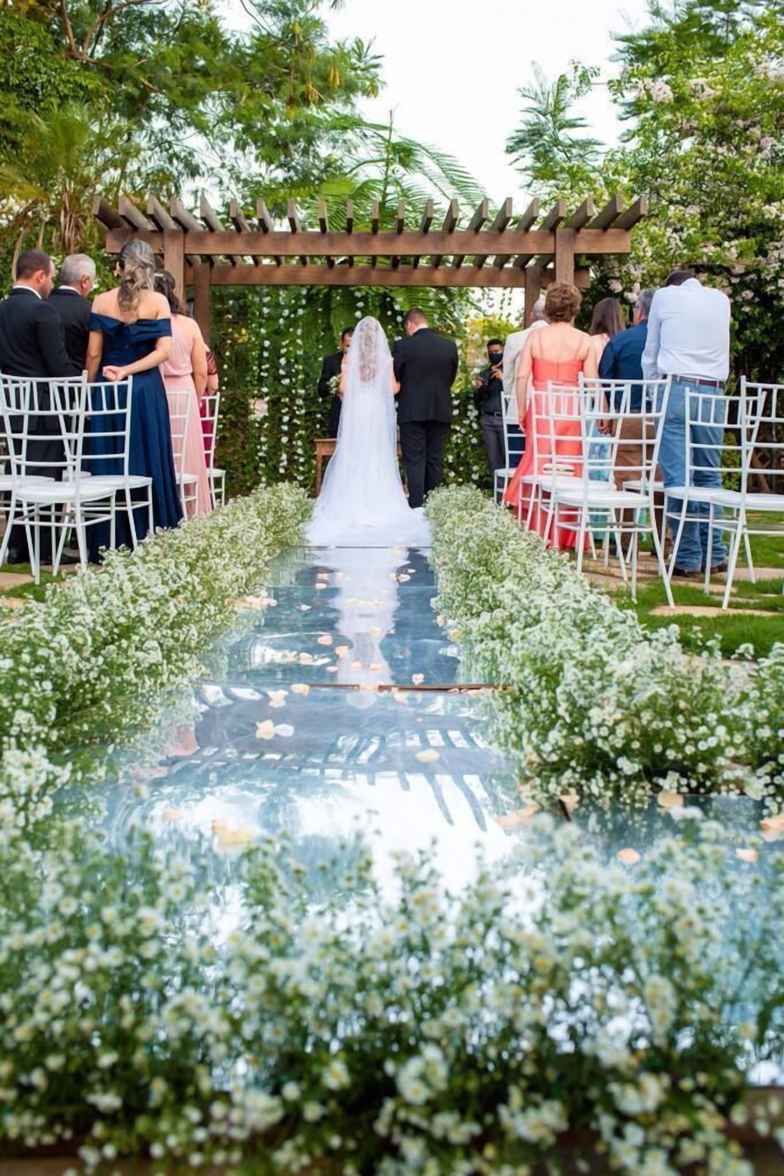 A wedding aisle with guests seated on white chairs, a wooden pergola, and a floral arrangement.