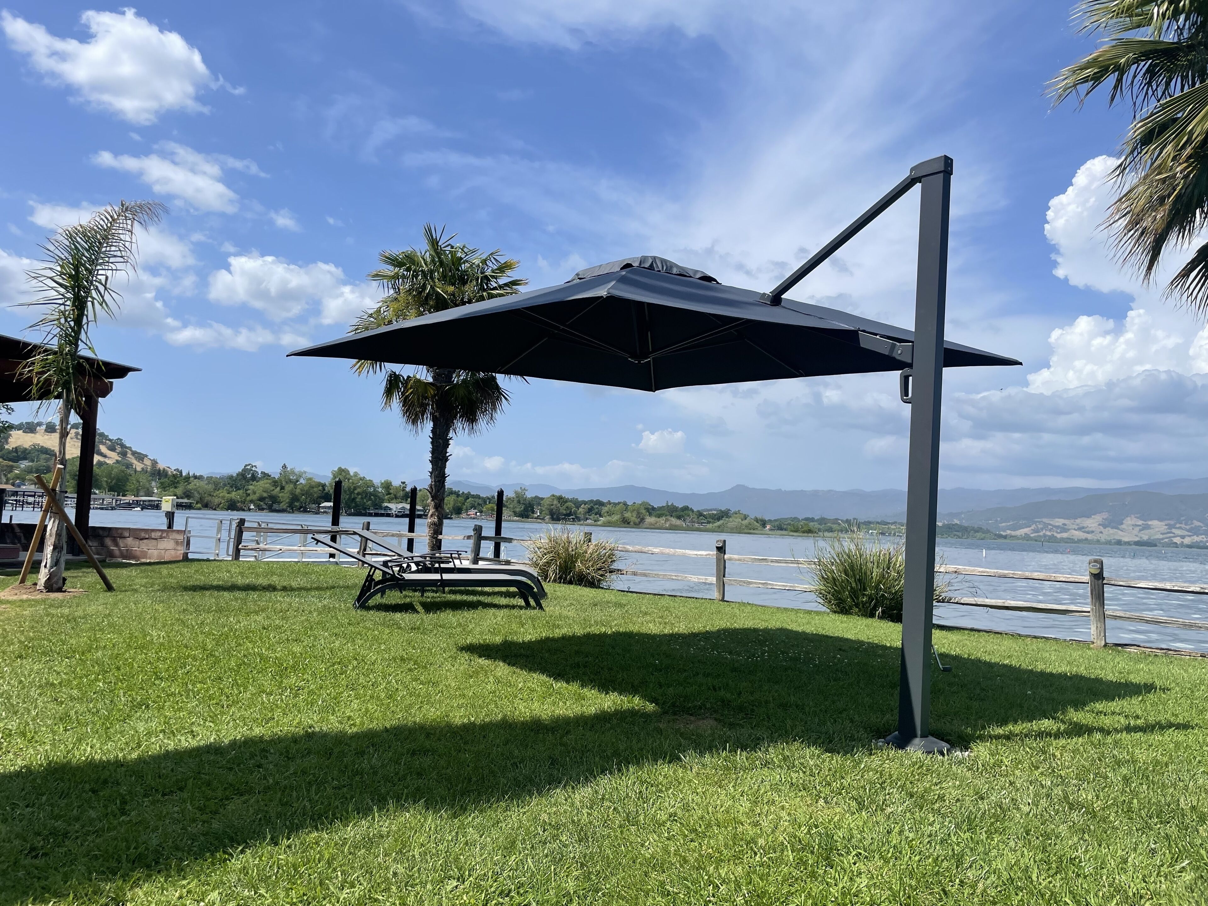 A grassy area with a lounge chair and umbrella, a wooden fence, and a view of a lake with mountains in the background.