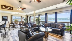 A spacious living area with a large sectional sofa, a glass coffee table, and a dining table with chairs. There are large windows offering a view of the outdoors.