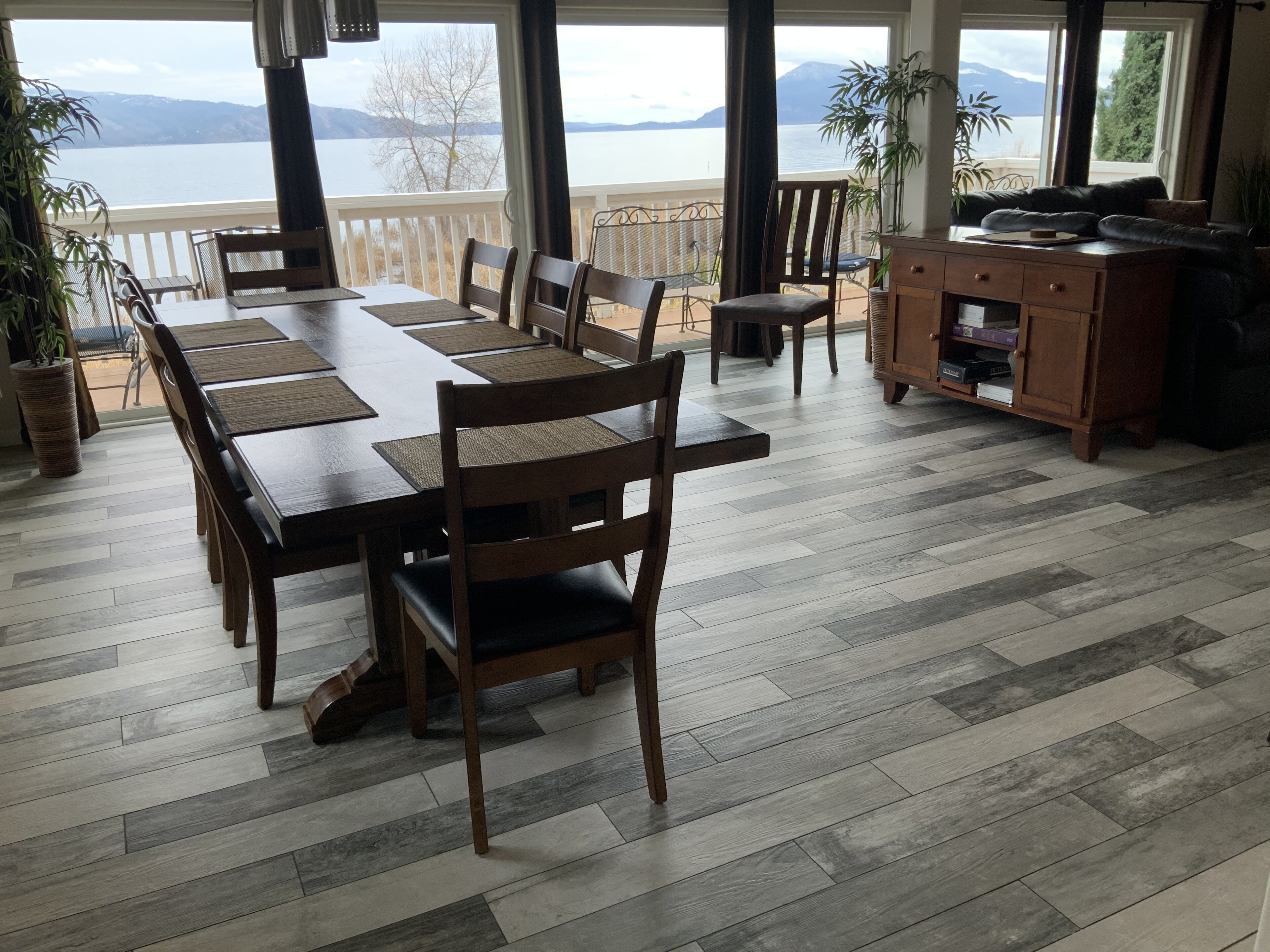 A dining area with a large wooden table and chairs, a kitchen area with a wooden cabinet, and a view of a lake through large windows.