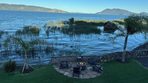 A patio with outdoor furniture and a fire pit, overlooking a lake with a dock and surrounded by grass and trees.