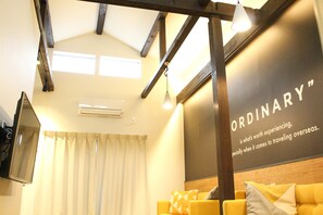 A hotel room with a high ceiling, exposed wooden beams, a large window with curtains, and a wall with the text "ORDINARY".
