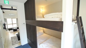 Habitación de hotel con literas, un escritorio y una silla. Hay una ventana con cortinas y una puerta con letrero de salida de emergencia.