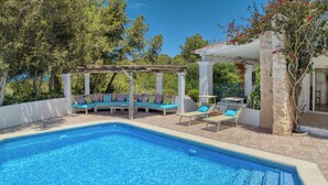 A swimming pool with a pergola and lounge chairs, surrounded by trees and a white building.