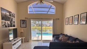 Ein großzügiges Wohnzimmer mit großem Fenster, Deckenventilator, Flachbildfernseher, einem dunkelgrauen Sofa und Blick auf einen Pool.