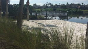 Ein sandiger Strandabschnitt mit einer Holzbrücke über dem Wasser, umgeben von Palmen und viel Grün.