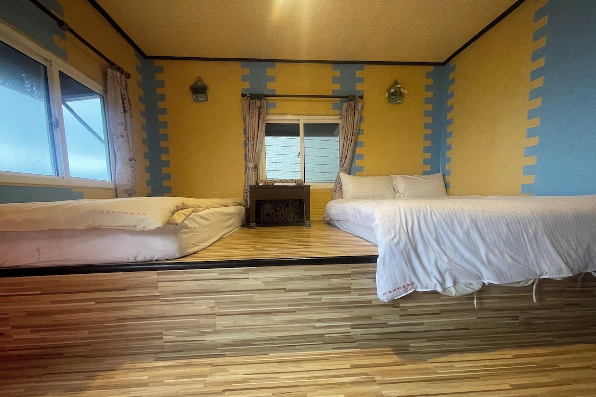 Une chambre avec deux lits, une fenêtre avec des rideaux, une petite table et un mur recouvert d’un rembourrage bleu et jaune.