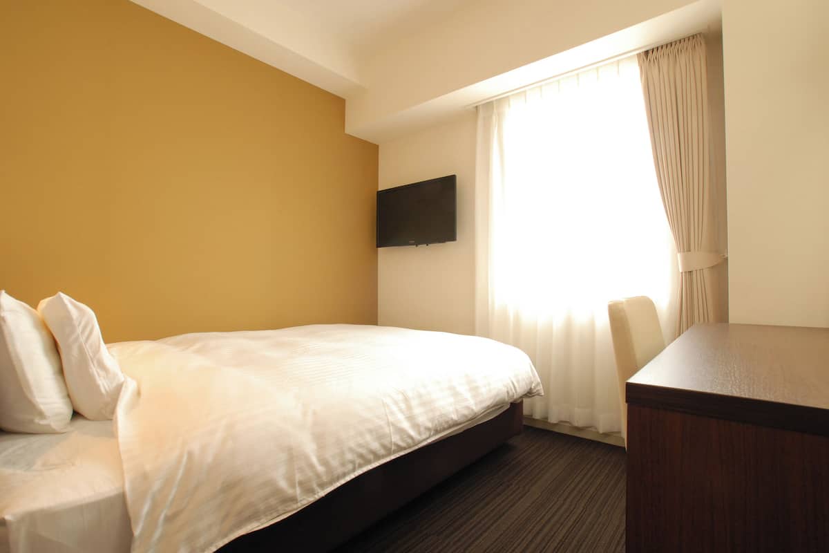 A hotel room with a bed, a desk, a flat-screen TV mounted on the wall, and a window with curtains.