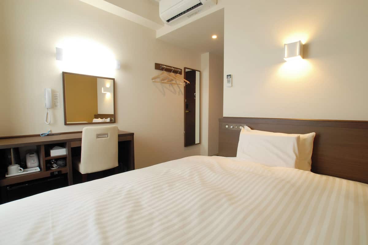 A hotel room with a bed, a desk with a chair, a mirror, a coat rack, and a wall-mounted phone.
