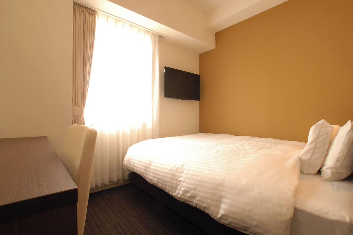 A hotel room with a bed, a desk, a chair, a flat-screen TV mounted on the wall, and a window with curtains.