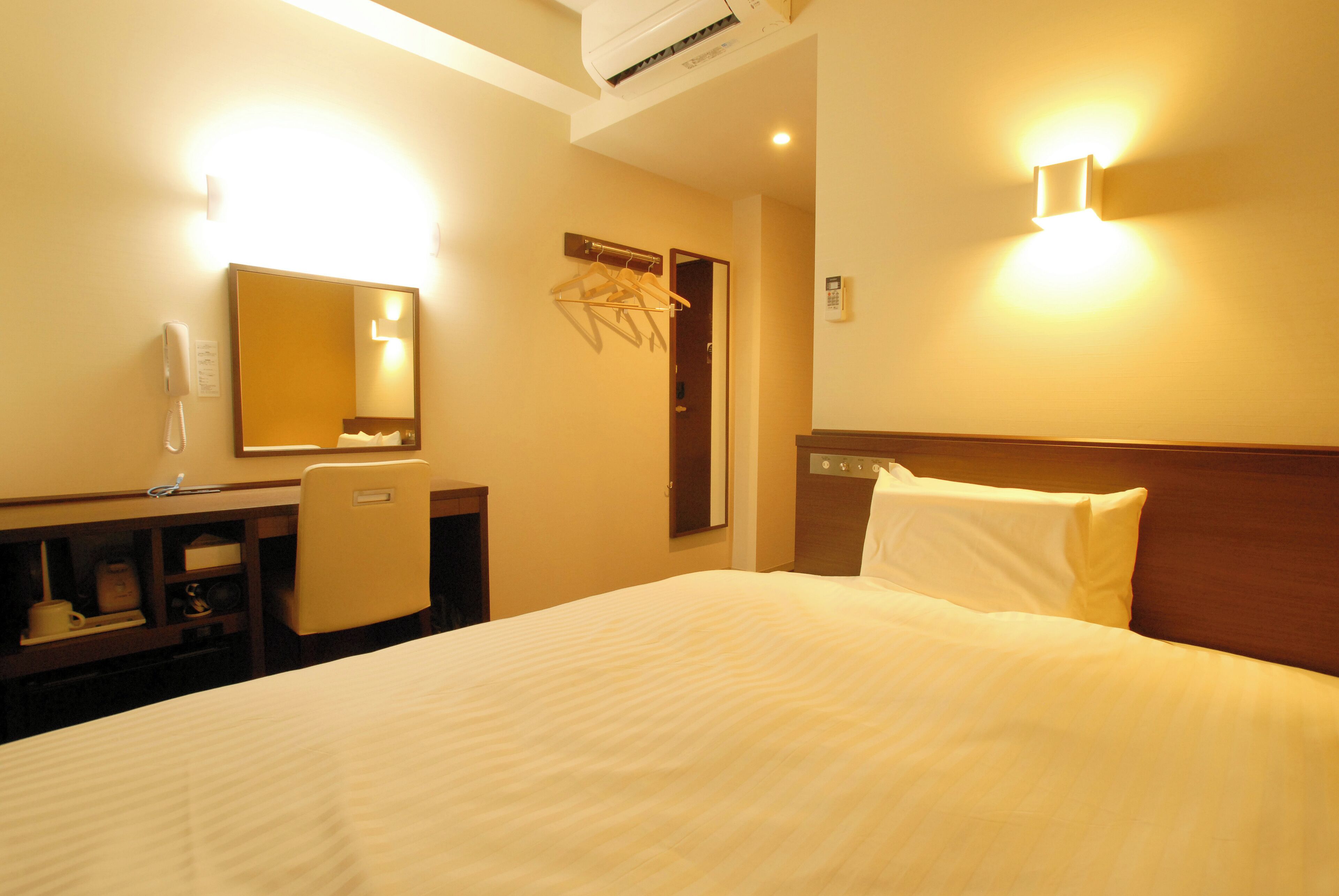 Habitación de hotel con cama, escritorio, silla, espejo, teléfono instalado en la pared y lámpara de pared.