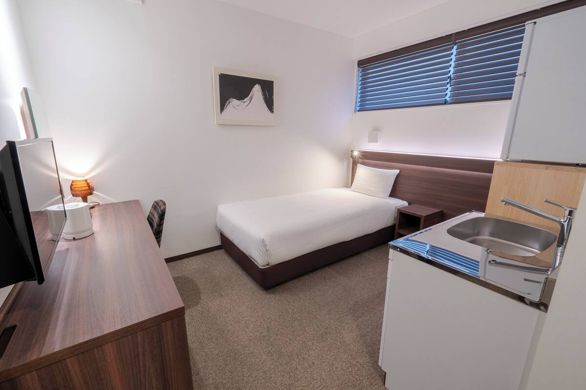 Ein Hotelzimmer mit einem Bett, einem Schreibtisch mit Computer, einem Waschbecken und einem Fenster mit Jalousien.