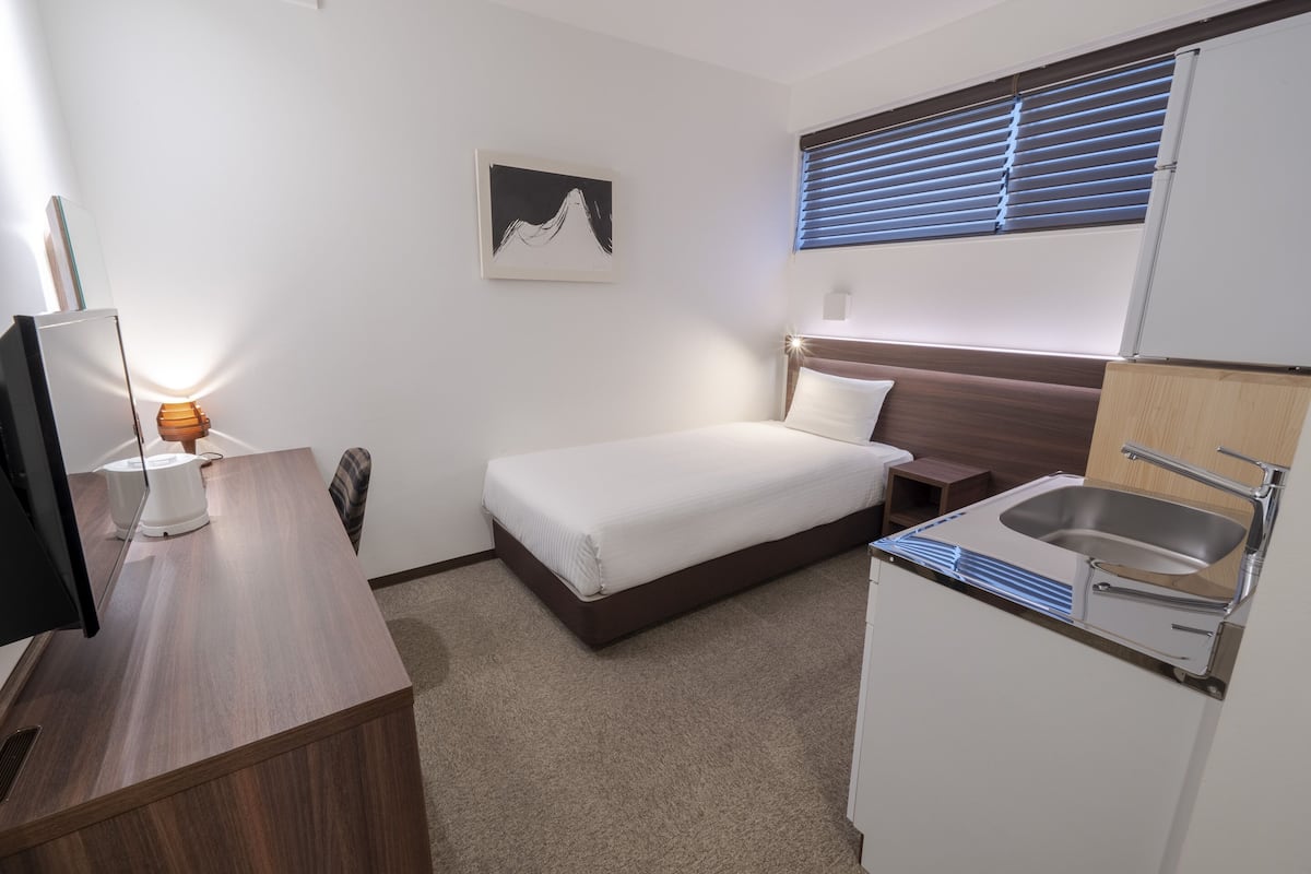 Ein Hotelzimmer mit einem Bett, einem Schreibtisch mit Computer, einem Waschbecken und einem Fenster mit Jalousien.
