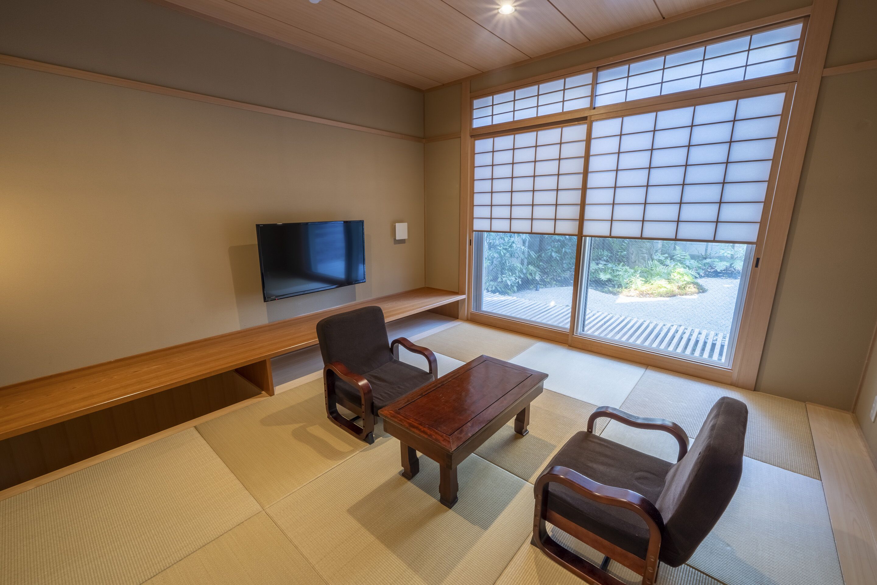 Une pièce de style japonais traditionnel, avec un sol recouvert de tatamis, une basse table en bois, deux chaises, un canapé et un téléviseur fixé au mur.