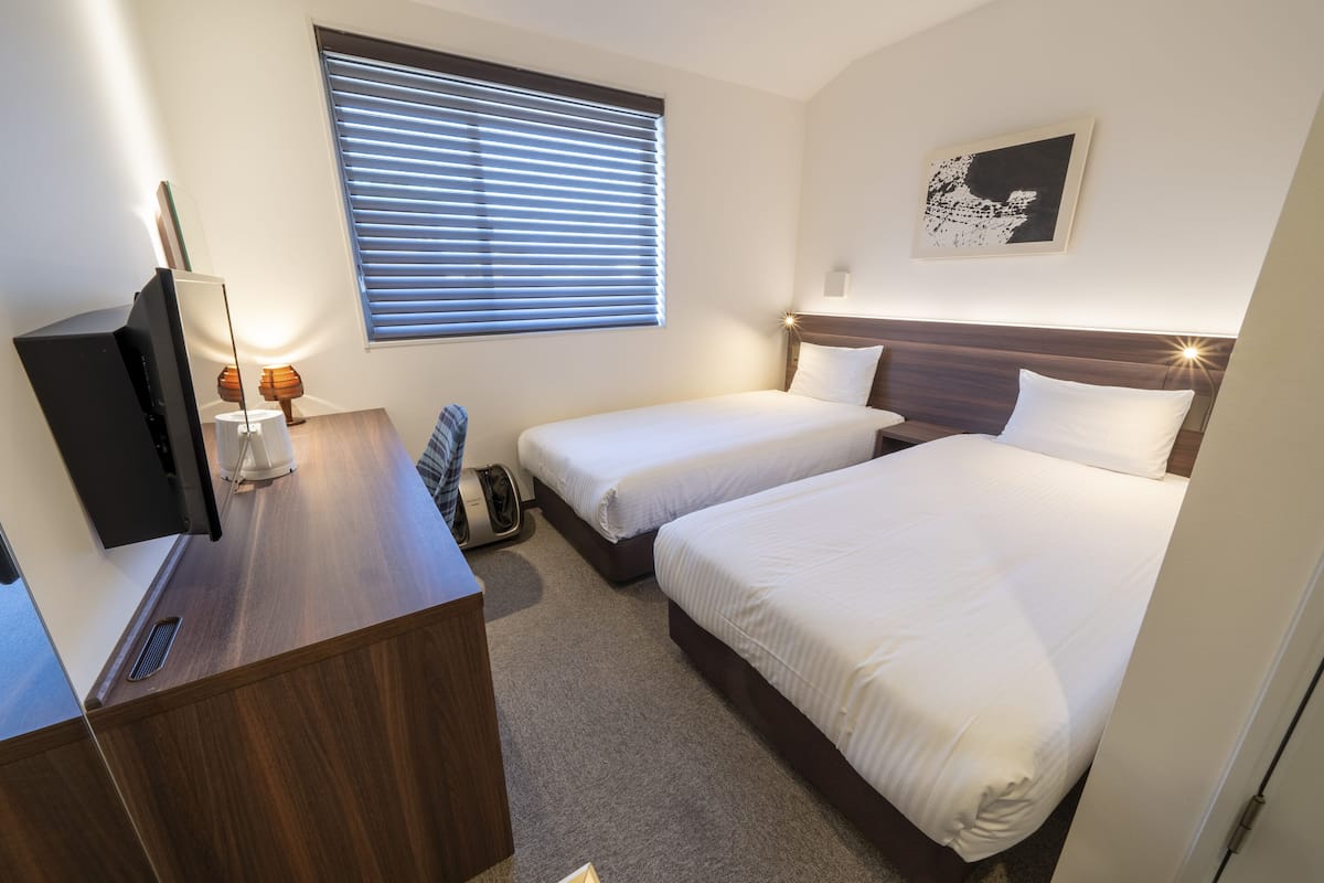Ein Hotelzimmer mit zwei Betten, einem Schreibtisch mit Fernseher, einem Fenster mit Jalousien und einem an der Wand hängenden Kunstwerk.