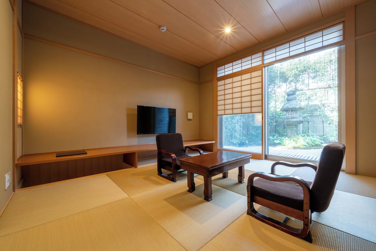 Ein traditioneller japanischer Raum mit Tatami-Boden, einem niedrigen Holztisch, zwei Stühlen, einem Fernseher und einem Fenster mit Blick auf Grünflächen.