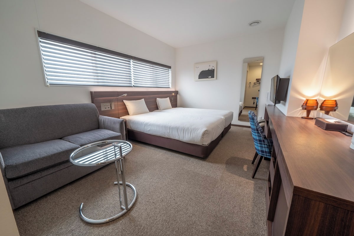 Ein modernes Hotelzimmer mit Bett, Sofa, kleinem Tisch, Schreibtisch mit Lampe und einem Fenster mit Jalousien.