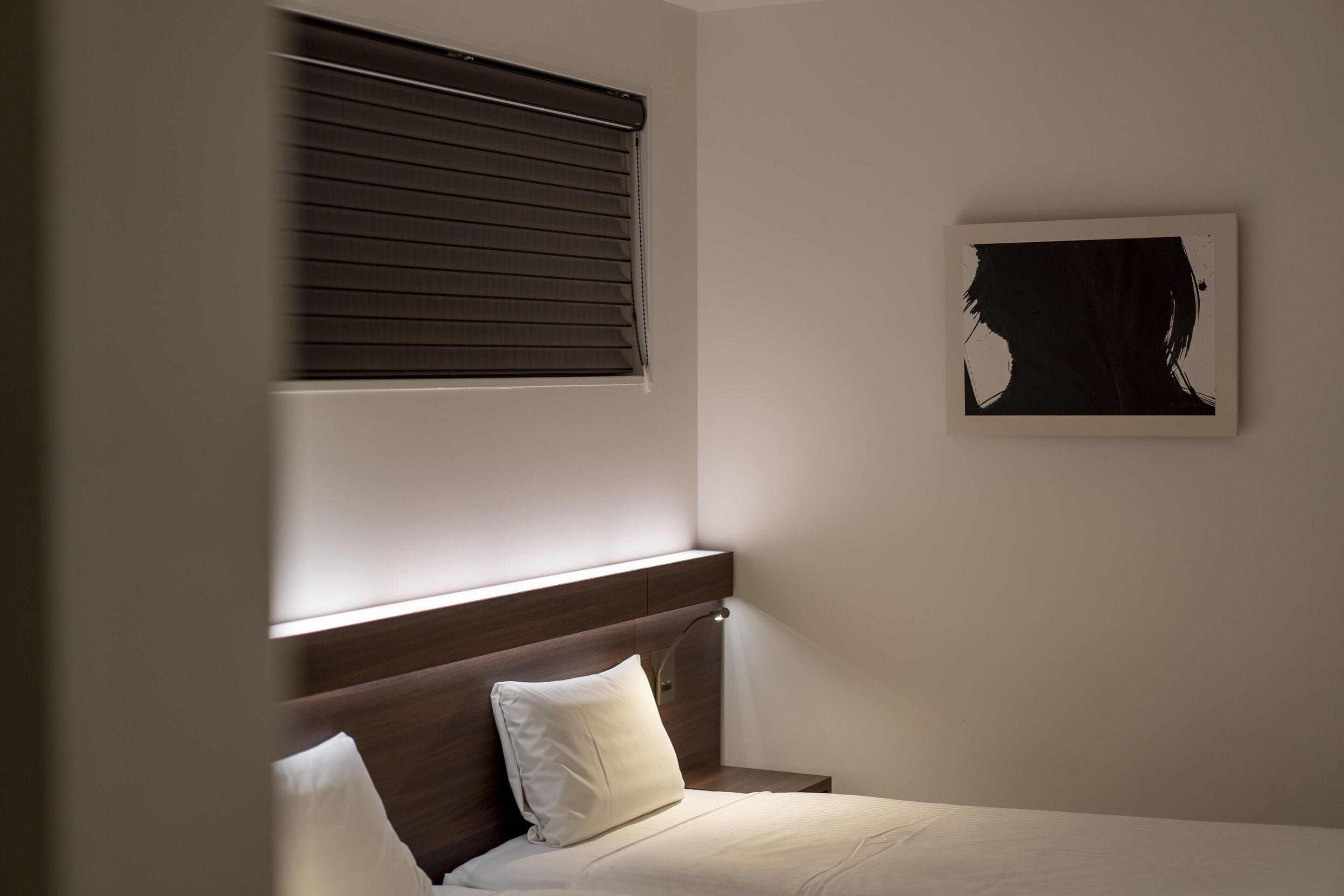 A hotel room with a bed, a nightstand, a wall-mounted lamp, a window with blinds, and a piece of abstract art.