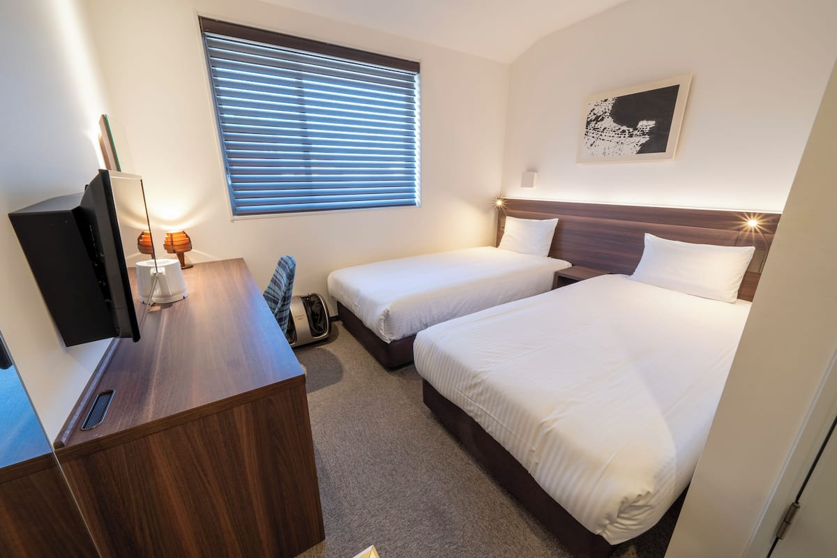 Ein Hotelzimmer mit zwei Betten, einem Schreibtisch mit Fernseher, einem Fenster mit Jalousien und einem an der Wand hängenden Kunstwerk.