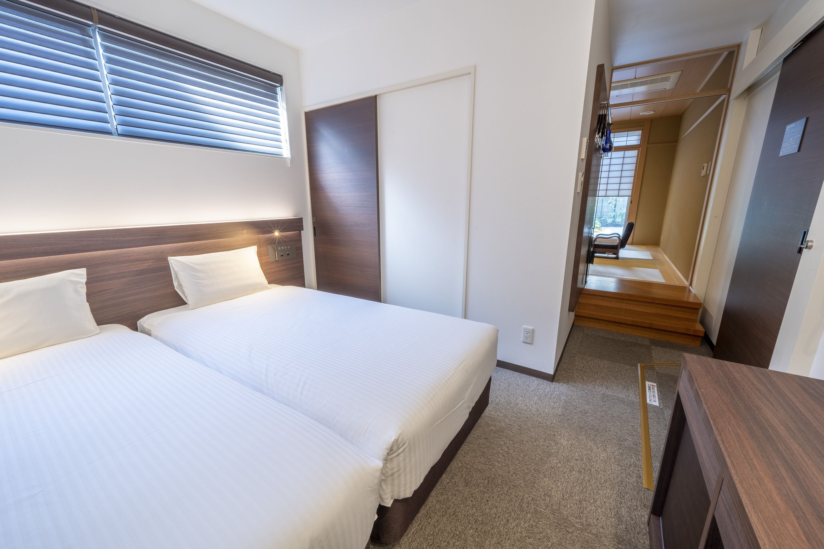 A hotel room with two beds, a wooden headboard, a sliding door, and a view of a wooden staircase.