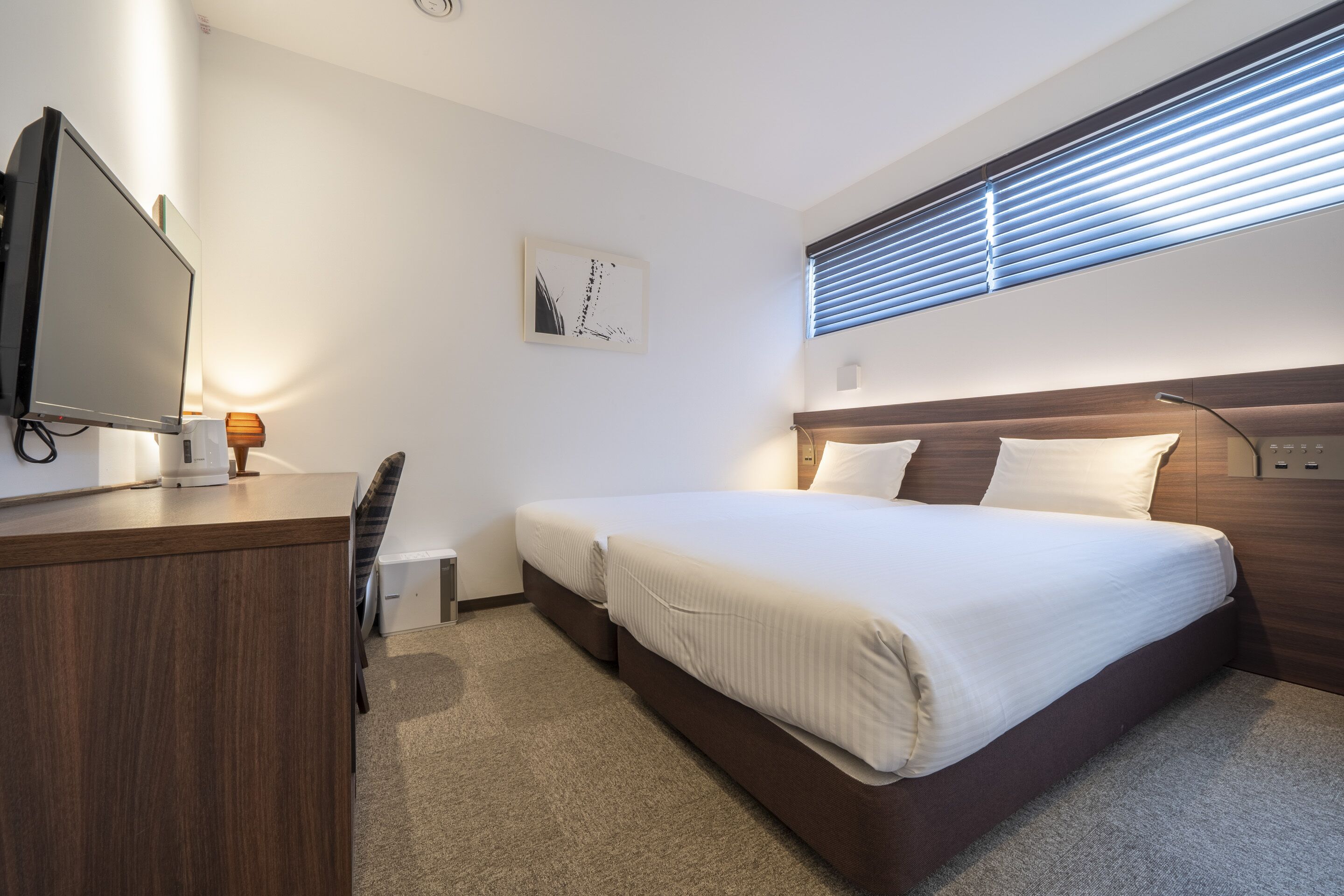 A hotel room with a bed, a desk with a computer, a chair, and a window with blinds.