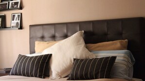 A neatly made bed with a tufted headboard, multiple pillows, and a plain wall with framed pictures.