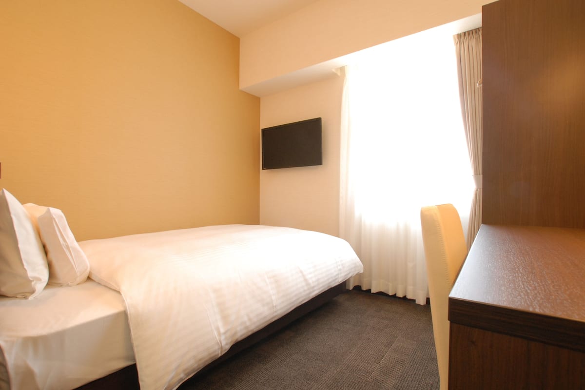 A hotel room with a single bed, a flat-screen TV mounted on the wall, and a window with curtains.