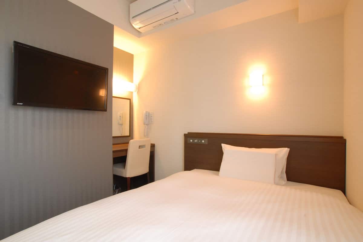 A hotel room with a bed, a desk, a chair, a flat-screen TV, and a wall-mounted light fixture.