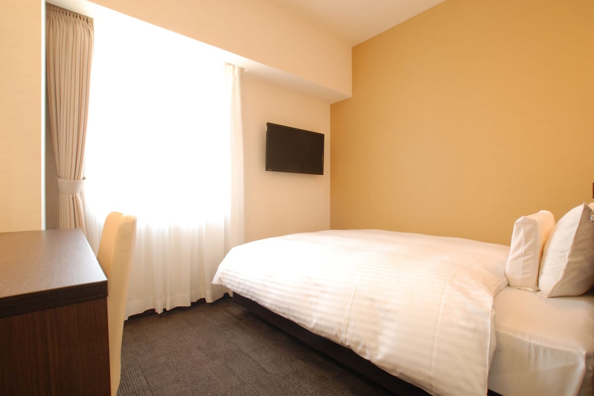 A hotel room with a bed, a desk, a chair, a flat-screen TV mounted on the wall, and a window with curtains.