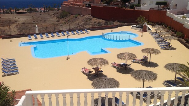 A pool area with sun loungers and umbrellas, surrounded by buildings and a view of the sea.