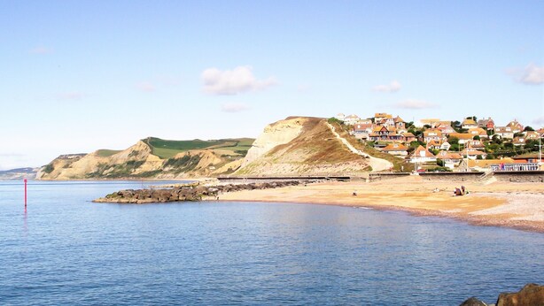 A coastal town with a rocky shoreline, a pier, and houses with a hillside backdrop.