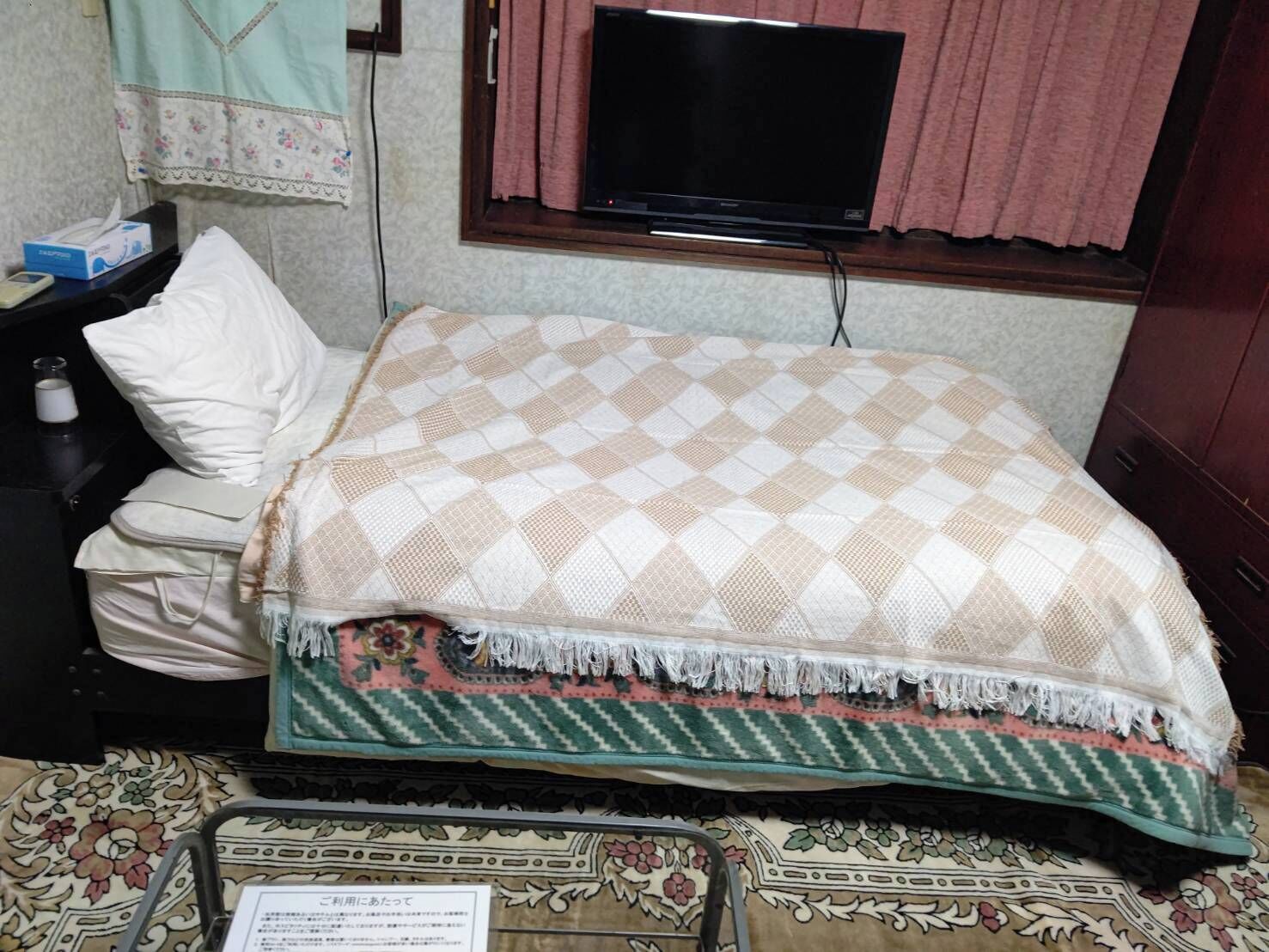 Uma cama com um cobertor xadrez, uma mesa de cabeceira com um estojo de lenços e uma televisão na parede.