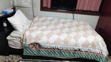 Uma cama com um cobertor xadrez, uma mesa de cabeceira com um estojo de lenços e uma televisão na parede.