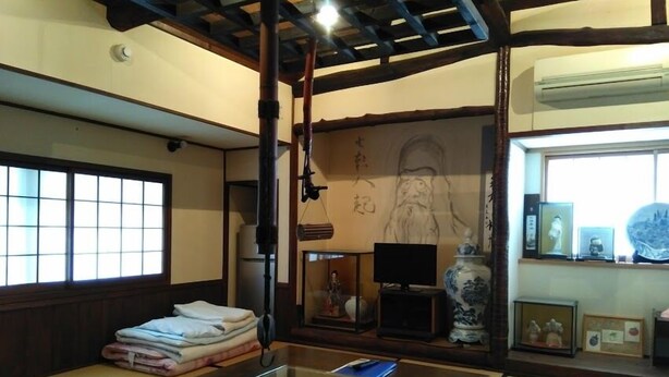 Una habitación tradicional japonesa con suelo de tatami, una mesa de vidrio, un televisor, un jarrón grande y un retrato enmarcado en la pared.