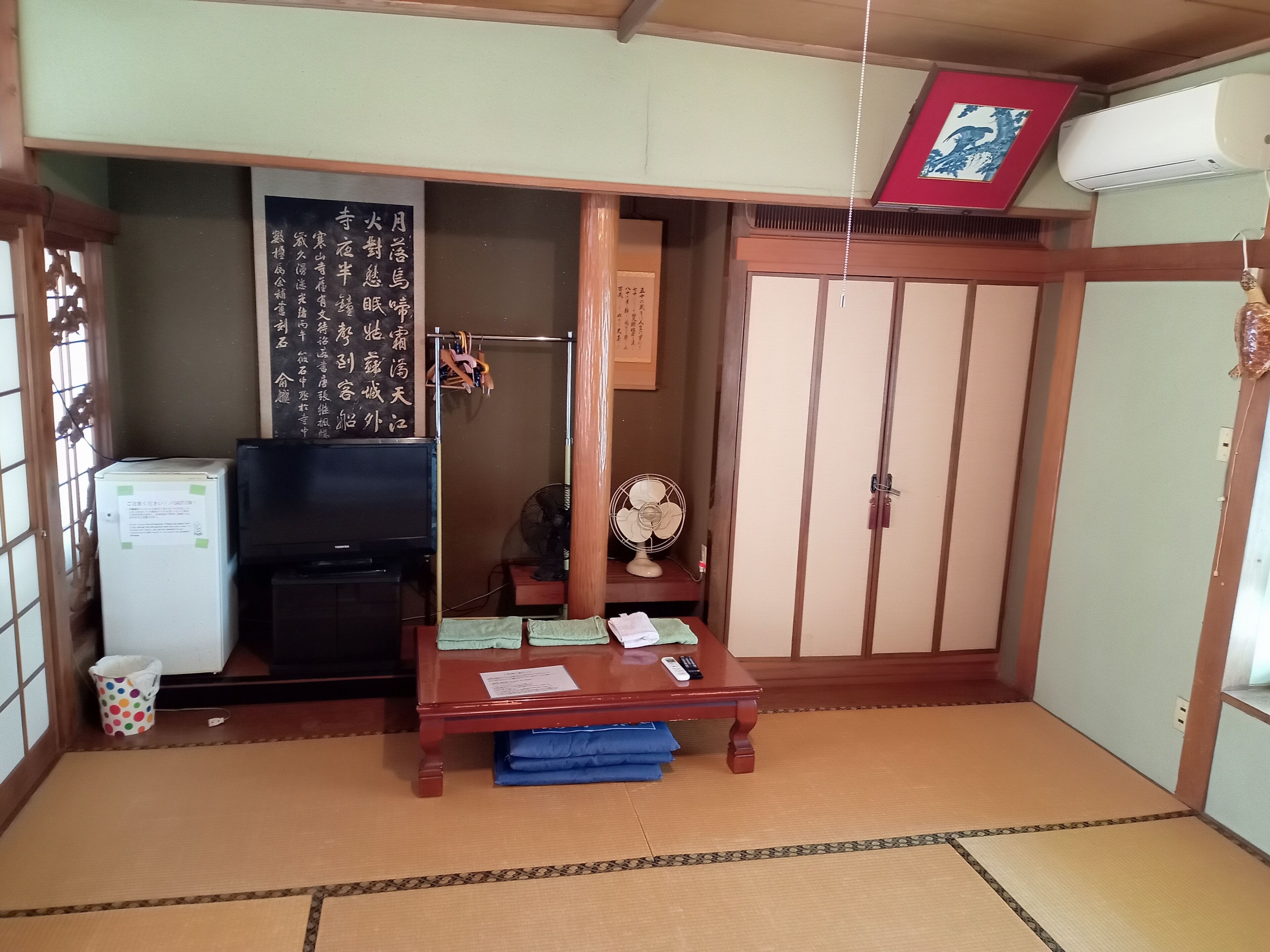 Ett traditionellt japanskt rum med tatamimattor på golvet, en tv, ett kylskåp, ett träbord med en fläkt och skjutdörrar.