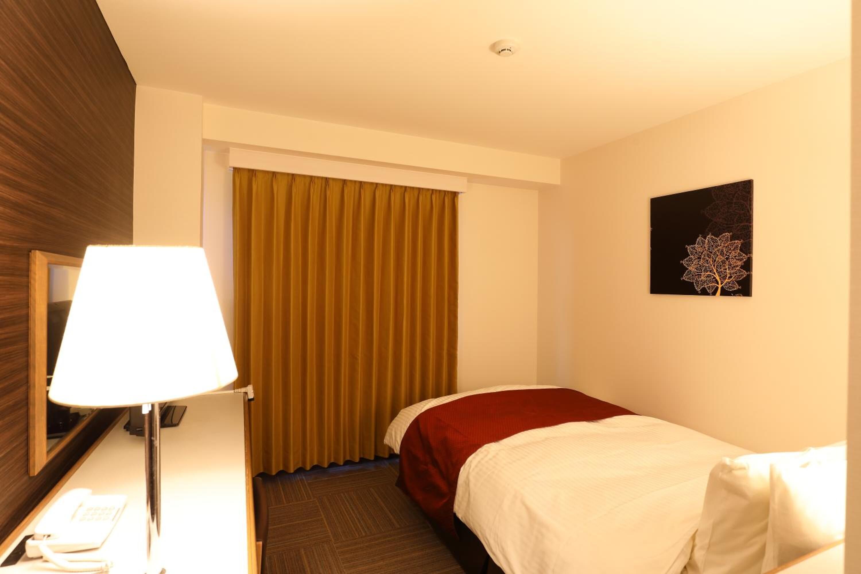 A hotel room with a bed, a desk with a lamp, a telephone, and a painting on the wall.