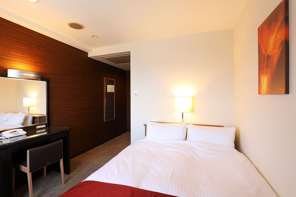 A hotel room with a bed, a desk with a chair, a mirror, and a wall-mounted lamp.