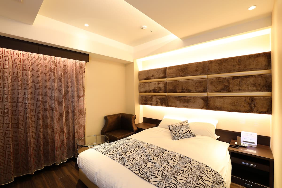 A modern hotel room with a large bed, a headboard with a textured design, a bedside table with a lamp, and a patterned curtain.