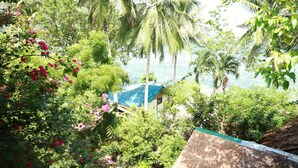 Um jardim tropical exuberante com palmeiras e plantas floridas, uma construção com teto de palha e vista para o mar.