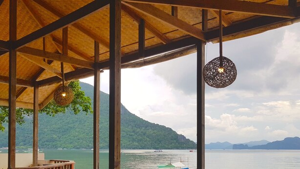 Uma área de estar coberta ao ar livre, com mobiliário de vime e luminárias pendentes, com vista para um corpo d'água calmo, barcos e um cenário montanhoso.