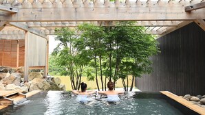 Una pérgola de madera sobre un área de aguas termales, con dos personas relajándose en el agua, rodeadas de rocas y árboles.