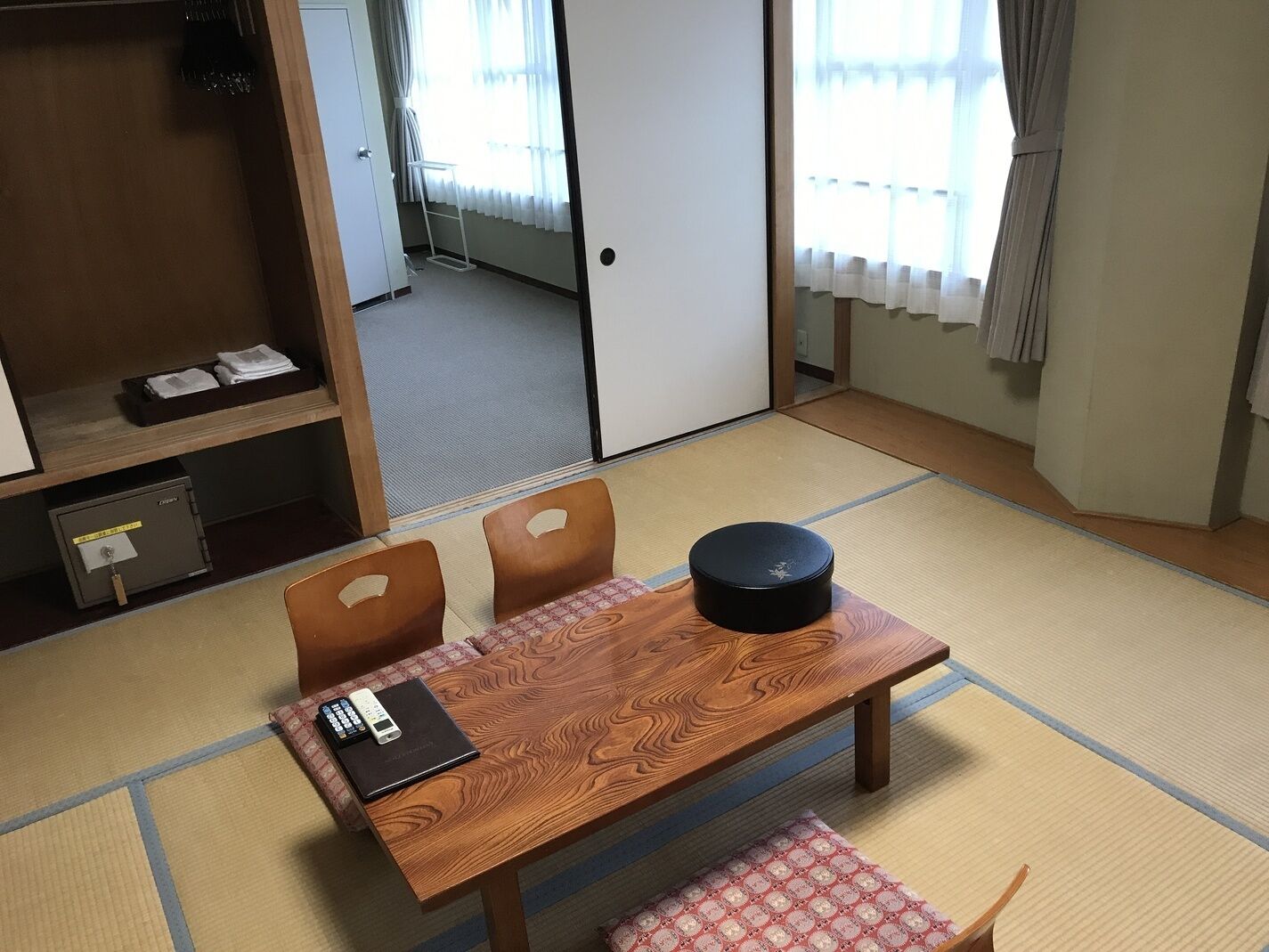 A traditional Japanese room with tatami flooring, a low wooden table with a black container, a chair, and a small shelf with a safe and folded clothes.