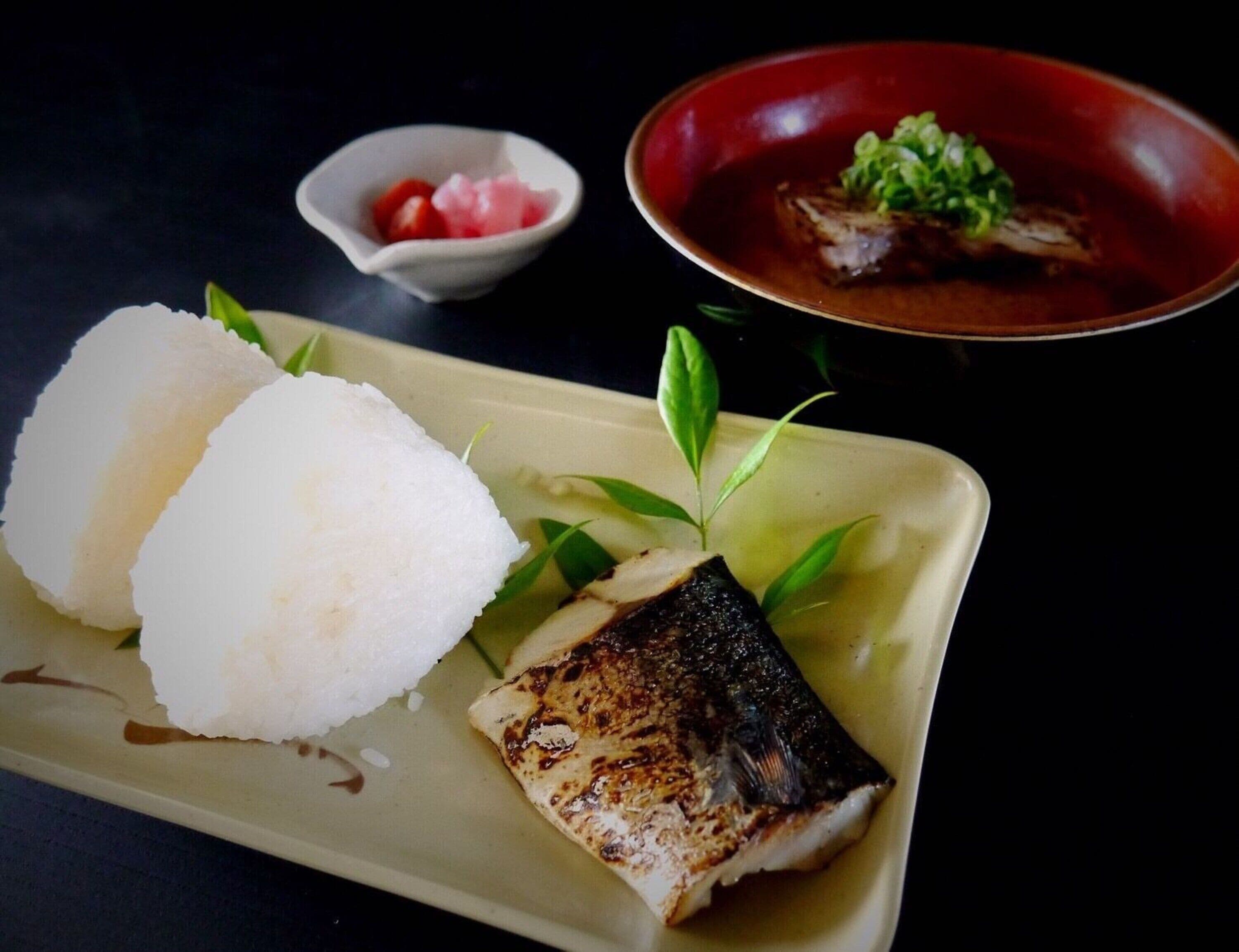 Ein Teller mit gegrilltem Fisch und weißem Reis, begleitet von einer kleinen Schale eingelegtes Gemüse und einer roten Schale Suppe.