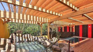 Una pérgola de madera sobre un área de hidromasaje con agua cristalina, rocas y un muro rojo y negro al fondo.