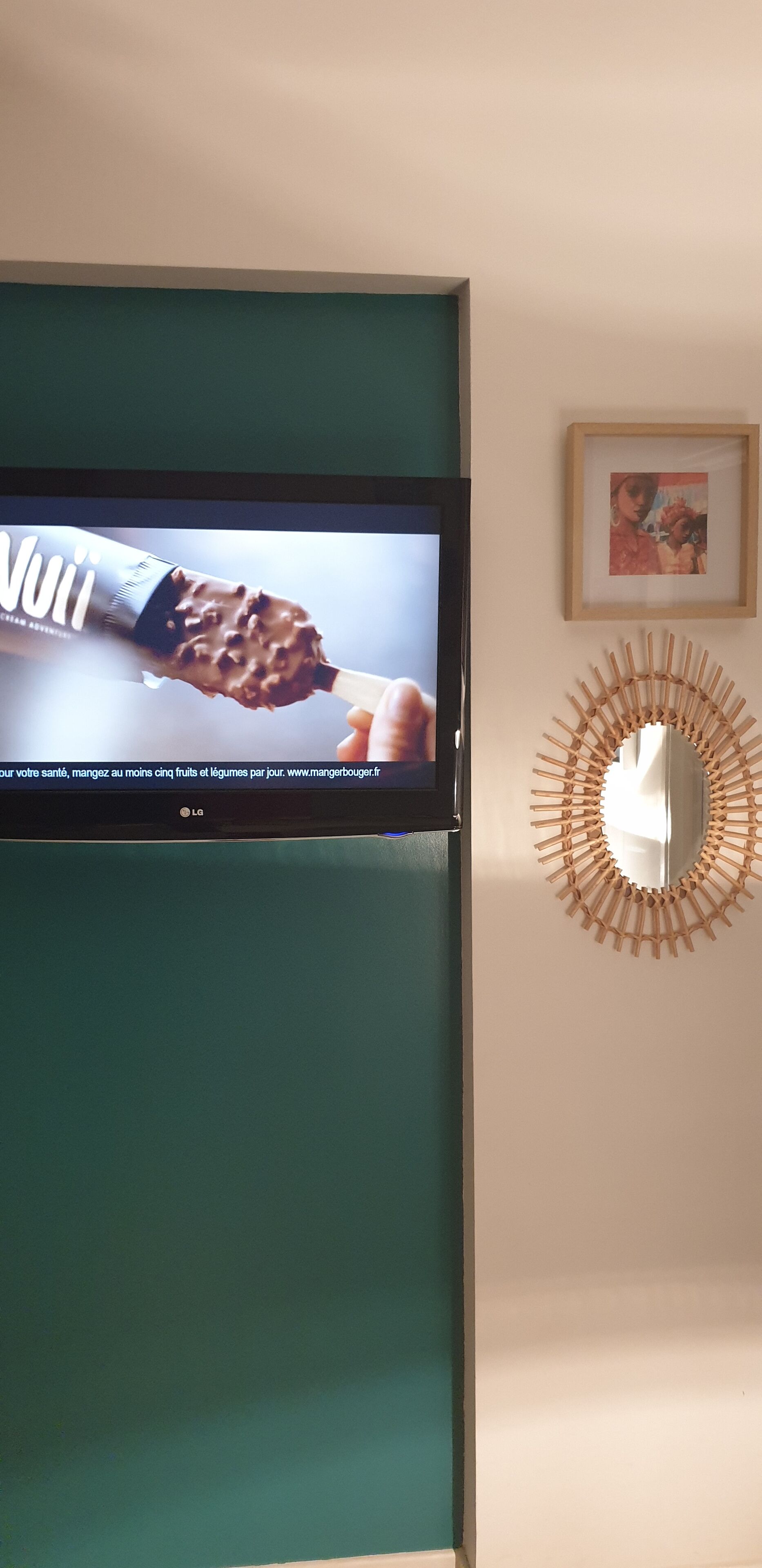Un televisor montado en una pared verde azulada, un cuadro enmarcado encima y un espejo de madera con diseño de sol.